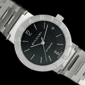 May include: A silver stainless steel wristwatch with a black face and silver hands. The watch face has a circular design with Roman numerals around the edge. The watch face also has the words "BVLGARI" and "AUTOMATIC" printed on it.
