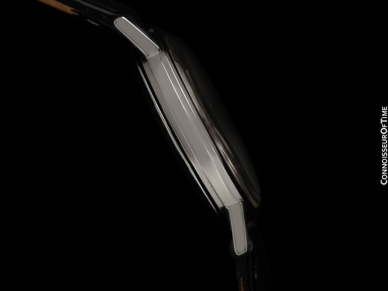 May include: Side view of a silver wristwatch with a black leather band. The watch has a rectangular face and a polished metal case. The image is set against a black background. The text "CONNOISSEUROFTIME" is visible.