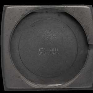 K&ouml;nnte beinhalten: Nahaufnahme der R&uuml;ckseite eines Uhrengeh&auml;uses mit kreisf&ouml;rmiger Mitte und quadratischem Au&szlig;enrahmen. Der Text "STAINLESS STEEL BACK" ist sichtbar, zusammen mit einem Logo. Das Geh&auml;use ist dunkelgrau.