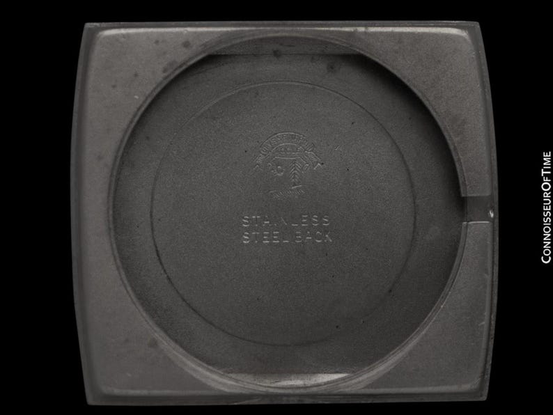 K&ouml;nnte beinhalten: Nahaufnahme der R&uuml;ckseite eines Uhrengeh&auml;uses mit kreisf&ouml;rmiger Mitte und quadratischem Au&szlig;enrahmen. Der Text "STAINLESS STEEL BACK" ist sichtbar, zusammen mit einem Logo. Das Geh&auml;use ist dunkelgrau.