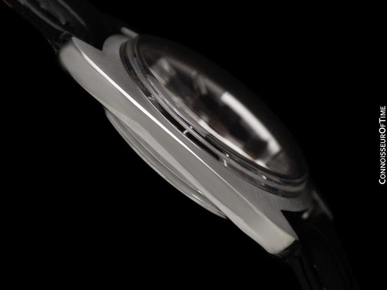 May include: Close-up of a silver wristwatch with a black leather band. The watch has a clear, domed crystal and a sleek, metallic case. The image is set against a black background, highlighting the watch's design. The text "CONNOISSEUROFTIME" is visible.