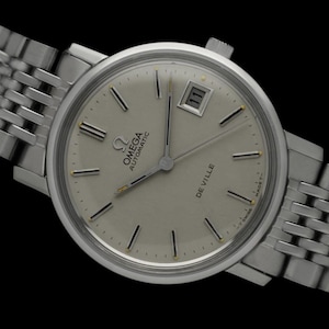 May include: A silver Omega De Ville Automatic wristwatch with a white face and black Roman numeral hour markers. The watch has a date window at the 3 o'clock position and a silver metal bracelet.