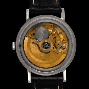 May include: Close-up of a silver wristwatch with a black leather band, showcasing the intricate gold-toned mechanical movement. The watch face is visible, revealing gears and other internal components. The watch is against a black background.