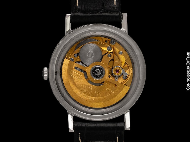 May include: Close-up of a silver wristwatch with a black leather band, showcasing the intricate gold-toned mechanical movement. The watch face is visible, revealing gears and other internal components. The watch is against a black background.