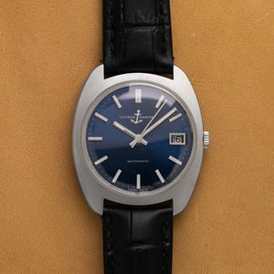 May include: A vintage Ulysse Nardin automatic wristwatch with a blue dial and silver hands. The watch has a date display at the 3 o'clock position and a black leather strap. The watch case is silver and has a unique shape.