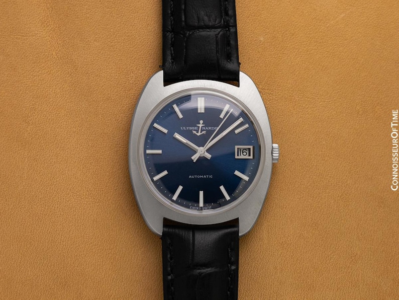 May include: A vintage Ulysse Nardin automatic wristwatch with a blue dial and silver hands. The watch has a date display at the 3 o'clock position and a black leather strap. The watch case is silver and has a unique shape.