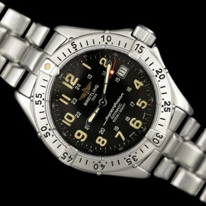 May include: A Breitling Superocean automatic wristwatch with a black dial and silver-tone stainless steel band. The watch features luminous hands and markers, a date display, and a rotating bezel. The watch is water-resistant to 1000m/3300ft.