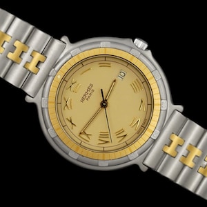 May include: A gold and silver two-tone wristwatch with a round face and Roman numeral hour markers. The watch face is cream colored with a gold-tone outer ring. The watch band is a two-tone metal link band with a gold H logo.