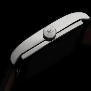 K&ouml;nnte beinhalten: Nahaufnahme einer silbernen Armbanduhr mit braunem Lederarmband. Die Uhr hat ein rechteckiges Zifferblatt und eine strukturierte Krone. Der Hintergrund ist schwarz. Der Text "CONNOISSEUROFTIME" ist auf der rechten Seite sichtbar.