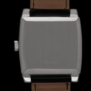 K&ouml;nnte beinhalten: Nahaufnahme einer silberfarbenen rechteckigen Armbanduhr mit braunem Lederarmband. Das Zifferblatt der Uhr ist nicht sichtbar. Auf der R&uuml;ckseite der Uhr ist die Nummer 4824607 eingraviert. Die Uhr ist vor schwarzem Hintergrund.