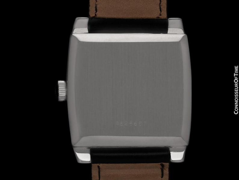 K&ouml;nnte beinhalten: Nahaufnahme einer silberfarbenen rechteckigen Armbanduhr mit braunem Lederarmband. Das Zifferblatt der Uhr ist nicht sichtbar. Auf der R&uuml;ckseite der Uhr ist die Nummer 4824607 eingraviert. Die Uhr ist vor schwarzem Hintergrund.