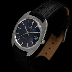 May include: A vintage Ulysse Nardin automatic watch with a dark blue dial and silver-tone case. The watch features a date window and is set on a black leather strap. The watch face has silver hands and markers. The watch is a classic timepiece.