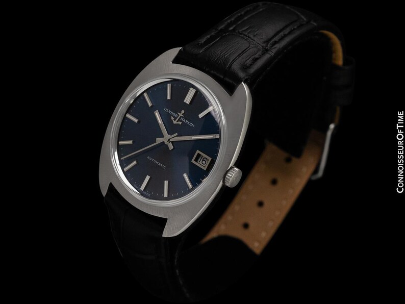 May include: A vintage Ulysse Nardin automatic watch with a dark blue dial and silver-tone case. The watch features a date window and is set on a black leather strap. The watch face has silver hands and markers. The watch is a classic timepiece.