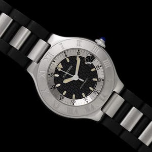 May include: A silver-toned Cartier watch with a black patterned dial and Roman numeral hour markers. The watch features a black rubber strap with silver-toned metal accents. The watch has a blue crown and the word "Cartier" is visible on the dial.