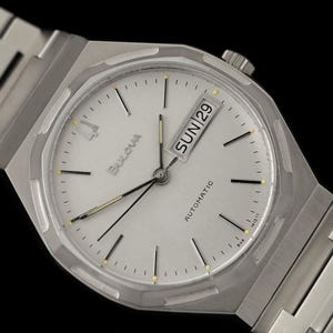 May include: A silver-toned Bulova automatic wristwatch with a white face. The watch features a day and date display, black hands, and a silver link band. The watch face has black and gold hour markers.