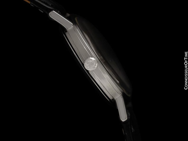 May include: Side view of a silver wristwatch with a black watch face and a black leather band. The watch has a round face and a small crown. The watch is set against a black background.