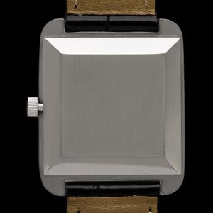 May include: A close-up of a rectangular silver watch with a black leather strap and a gold-colored underside. The watch has a silver crown and a brushed metal finish. The watch is set against a black background.