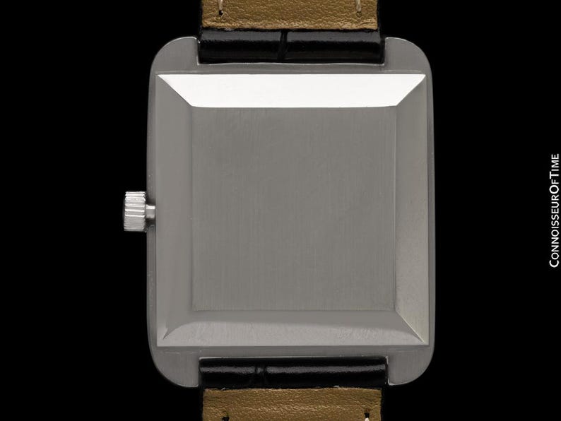 May include: A close-up of a rectangular silver watch with a black leather strap and a gold-colored underside. The watch has a silver crown and a brushed metal finish. The watch is set against a black background.