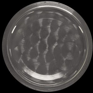 May include: A circular, metallic watch component with a brushed, swirling pattern. The component has a raised, rounded edge and a dark, reflective surface. The image is set against a black background, with the text "CONNOISSEUROFTIME" visible on the right side.