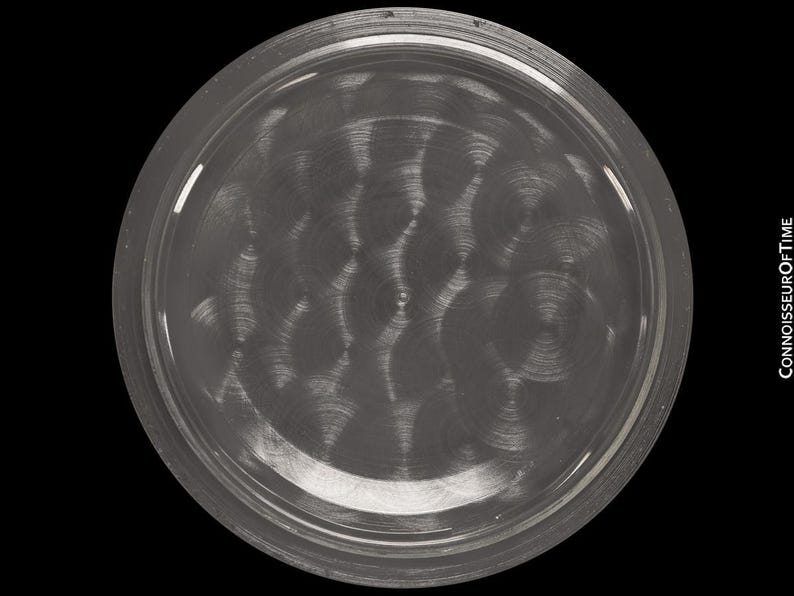 May include: A circular, metallic watch component with a brushed, swirling pattern. The component has a raised, rounded edge and a dark, reflective surface. The image is set against a black background, with the text "CONNOISSEUROFTIME" visible on the right side.