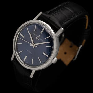 May include: A silver-toned wristwatch with a dark blue face and a black leather band. The watch face features silver hands and markers, with the text "ULYSSES NARDIN" and "AUTOMATIC". The band is textured with a crocodile pattern.