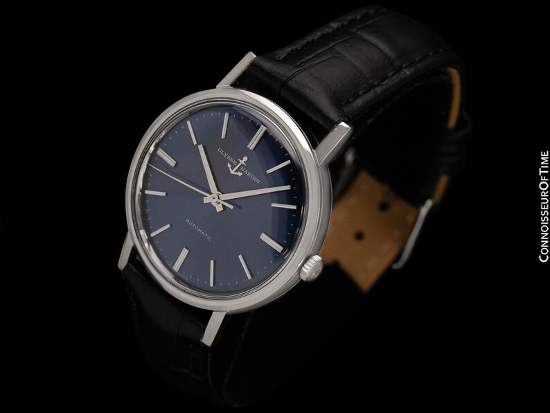 May include: A silver-toned wristwatch with a dark blue face and a black leather band. The watch face features silver hands and markers, with the text "ULYSSES NARDIN" and "AUTOMATIC". The band is textured with a crocodile pattern.