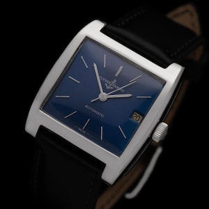 K&ouml;nnte beinhalten: Eine rechteckige Armbanduhr in Silber mit einem tiefblauen Zifferblatt und einem schwarzen Lederarmband. Das Zifferblatt hat silberne Zeiger und Markierungen mit dem Text "Ulysse Nardin" und "Automatic". Ein Datumsfenster zeigt die Zahl 20.
