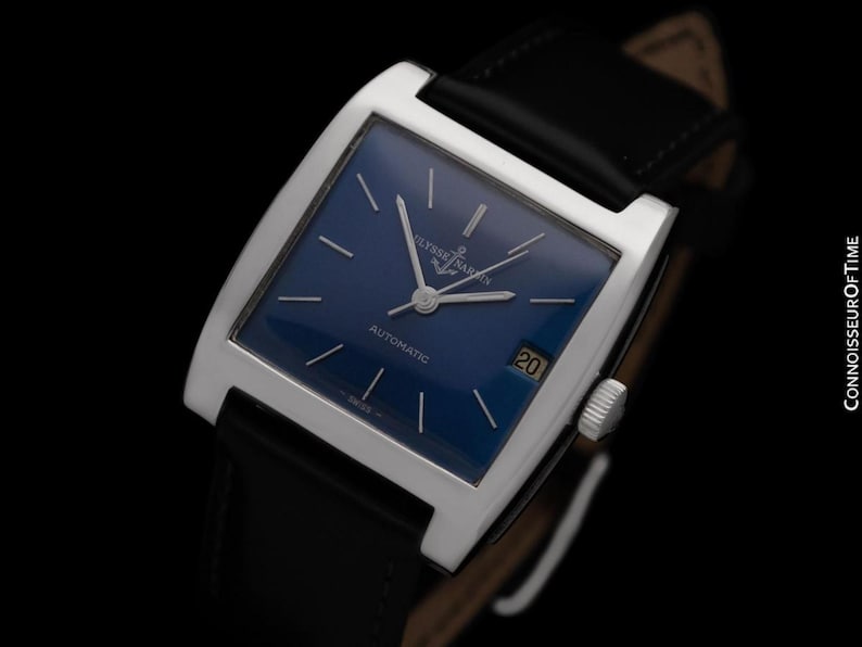 K&ouml;nnte beinhalten: Eine rechteckige Armbanduhr in Silber mit einem tiefblauen Zifferblatt und einem schwarzen Lederarmband. Das Zifferblatt hat silberne Zeiger und Markierungen mit dem Text "Ulysse Nardin" und "Automatic". Ein Datumsfenster zeigt die Zahl 20.