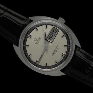 May include: A silver Omega Seamaster Cosmic automatic wristwatch with a white dial, black hands, and a day-date window. The watch has a black leather band.