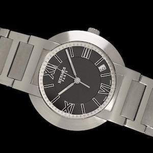 May include: A silver-toned wristwatch with a black face featuring Roman numerals and the brand name "Hermes Paris." The watch has a rectangular link band and a date display at the 3 o'clock position. The background is black.
