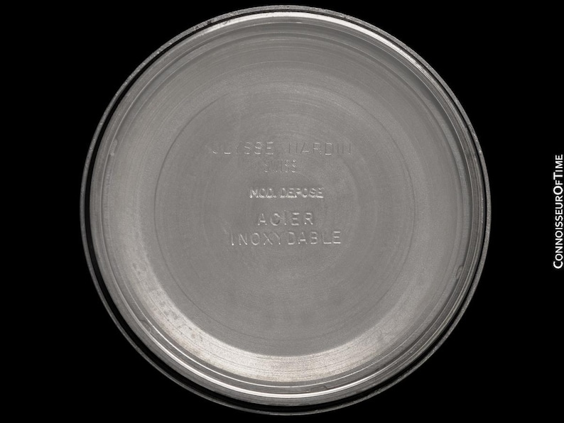 May include: A close-up of the back of a silver-colored watch. The watch back has the words "ULYSSE NARDIN" and other text in a circular pattern. The watch back is made of stainless steel.