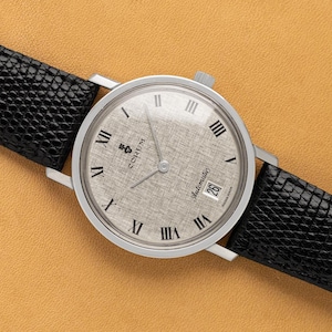 May include: A silver-toned Corum automatic wristwatch with a textured silver dial and black leather band. The watch face features Roman numeral hour markers and a date display at the 3 o'clock position. The black leather band has a textured pattern.