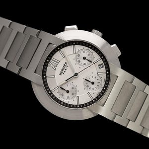 May include: A silver-toned wristwatch with a white face, black accents, and a date display. The watch features multiple sub-dials and a stainless steel band. The watch face displays the brand name "Hermes Paris".