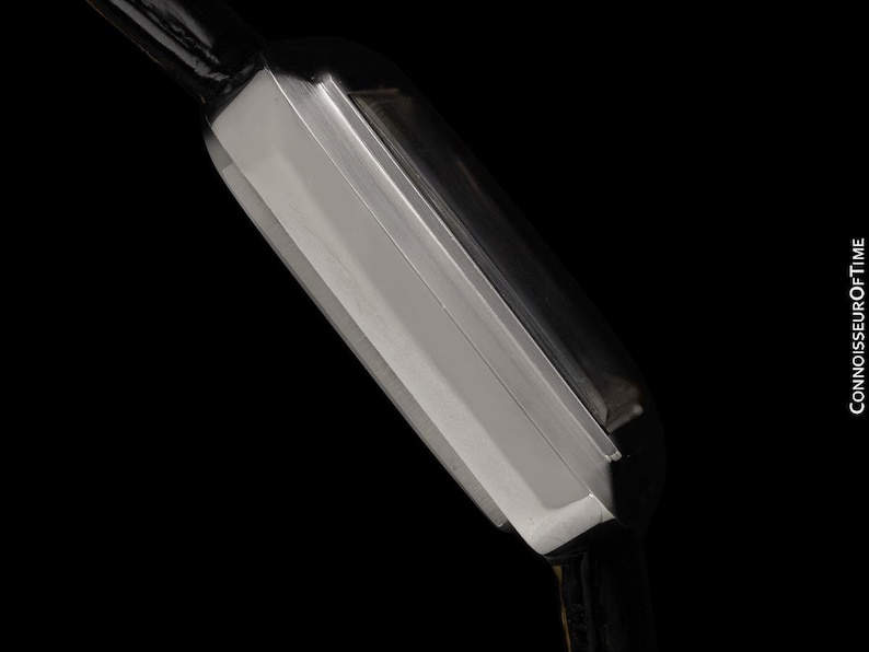 May include: Close-up of a rectangular silver wristwatch with a black leather strap. The watch has a sleek, modern design with a polished metal finish. The background is solid black, highlighting the watch's details.