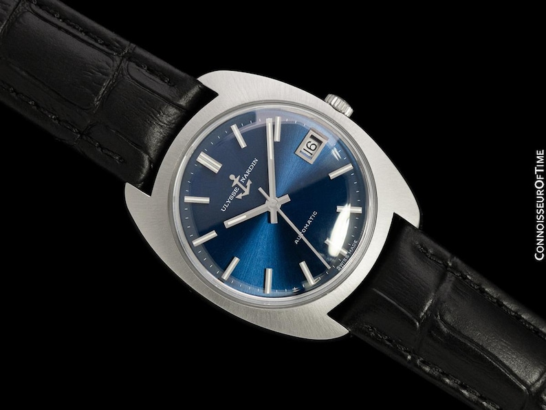 May include: A stainless steel Ulysse Nardin automatic watch with a blue dial and a black leather band. The watch features a date display at the 3 o'clock position and silver hour markers. The watch is set against a black background.
