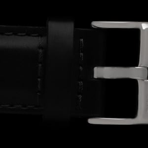K&ouml;nnte beinhalten: Nahaufnahme eines schwarzen Lederarmbands mit einer silberfarbenen Schnalle. Die Schnalle ist rechteckig mit abgerundeten Kanten und den Buchstaben "IWB" auf der Seite. Das Armband ist mit schwarzem Faden gen&auml;ht.