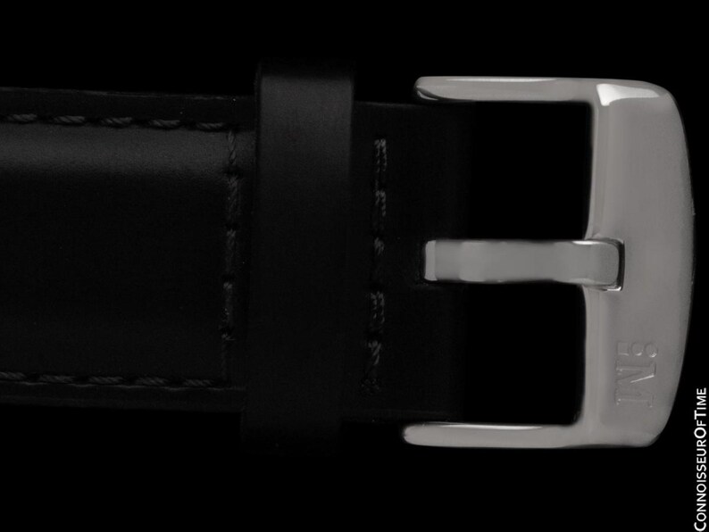 K&ouml;nnte beinhalten: Nahaufnahme eines schwarzen Lederarmbands mit einer silberfarbenen Schnalle. Die Schnalle ist rechteckig mit abgerundeten Kanten und den Buchstaben "IWB" auf der Seite. Das Armband ist mit schwarzem Faden gen&auml;ht.