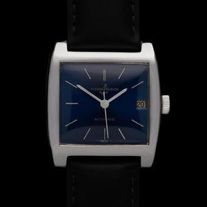 K&ouml;nnte beinhalten: Eine silberfarbene rechteckige Uhr mit dunkelblauem Zifferblatt und schwarzem Lederarmband. Das Zifferblatt hat silberne Zeiger und Markierungen, ein Datumsfenster bei 3 Uhr und den Text "AUTOMATIC". Die Uhr ist vor schwarzem Hintergrund.