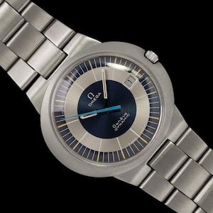 May include: A silver-toned Omega Genève Dynamic wristwatch with a blue and silver face. The watch features a date display at the 3 o'clock position and a silver metal band. The watch hands are blue and the watch face has the Omega logo.