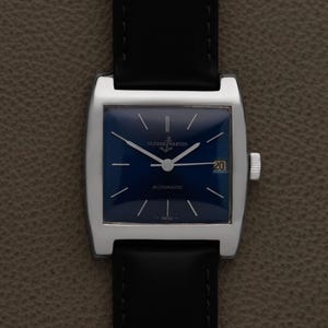 K&ouml;nnte beinhalten: Eine Vintage-Armbanduhr mit einem quadratischen, silberfarbenen Geh&auml;use und einem tiefblauen Zifferblatt. Die Uhr hat ein schwarzes Lederarmband und eine Datumsanzeige bei 3 Uhr. Das Zifferblatt hat silberfarbene Zeiger und Markierungen.