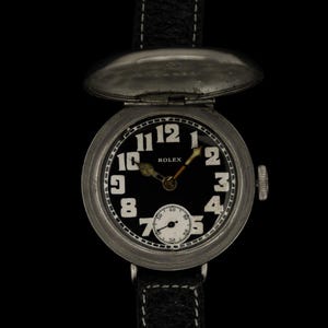 Puede incluir: Un reloj Rolex vintage con esfera negra y números blancos. El reloj tiene una caja plateada y una correa de cuero negro. La esfera incluye una subesfera más pequeña y el logotipo de Rolex. El reloj está sobre un fondo negro.