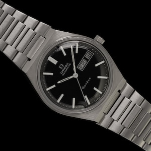 May include: A silver Omega Genève automatic wristwatch with a black dial. The watch has a date display and a stainless steel strap. The watch hands and hour markers are silver. The watch is set against a black background.