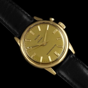 May include: A gold-toned Vulcain Cricket wristwatch with a black leather strap. The watch face is gold with rectangular hour markers and the words "Vulcain Cricket" and "Waterprotected". The watch has a textured crown.