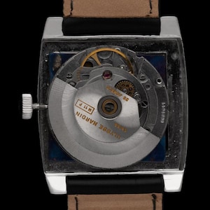 K&ouml;nnte beinhalten: Nahaufnahme einer rechteckigen silbernen Armbanduhr mit braunem Lederarmband. Das transparente Zifferblatt zeigt die komplizierten Innenmechanismen, einschlie&szlig;lich Zahnr&auml;dern und einem Rotor. Die Uhr ist mit "Ulysse Nardin" beschriftet.