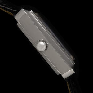 May include: A side view of a rectangular silver wristwatch with a black leather band. The watch has a white button on the side and a black face. The watch is set against a black background.
