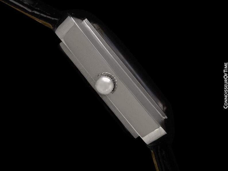May include: A side view of a rectangular silver wristwatch with a black leather band. The watch has a white button on the side and a black face. The watch is set against a black background.