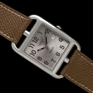 May include: A silver-toned rectangular watch with a light brown leather strap. The watch face is silver with black numerals and hands. The watch face reads "HERMES PARIS automatic SWISS MADE". The date is displayed at the 3 o'clock position.