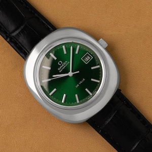 May include: A vintage Omega De Ville automatic watch with a green dial and silver case. The watch has a date window at the 3 o'clock position and is set on a black leather strap. The watch is displayed on a tan surface.