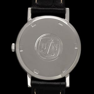 May include: Close-up of the back of a silver wristwatch with a black leather band. The watch face features a circular emblem with the letters "UN" and an anchor. The watch is set against a black background.