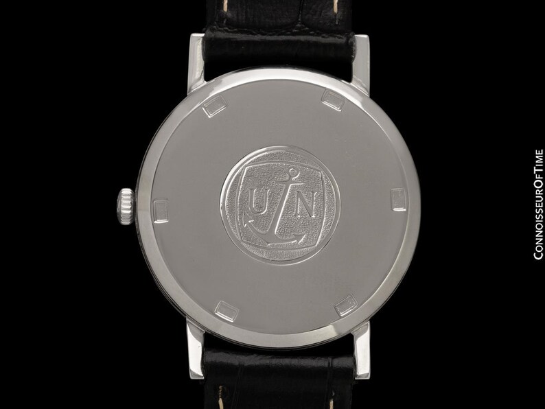 May include: Close-up of the back of a silver wristwatch with a black leather band. The watch face features a circular emblem with the letters "UN" and an anchor. The watch is set against a black background.